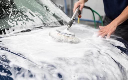 Operario aplicando espuma densa sobre capó de coche durante lavado exterior con agua a presión y cepillos suaves